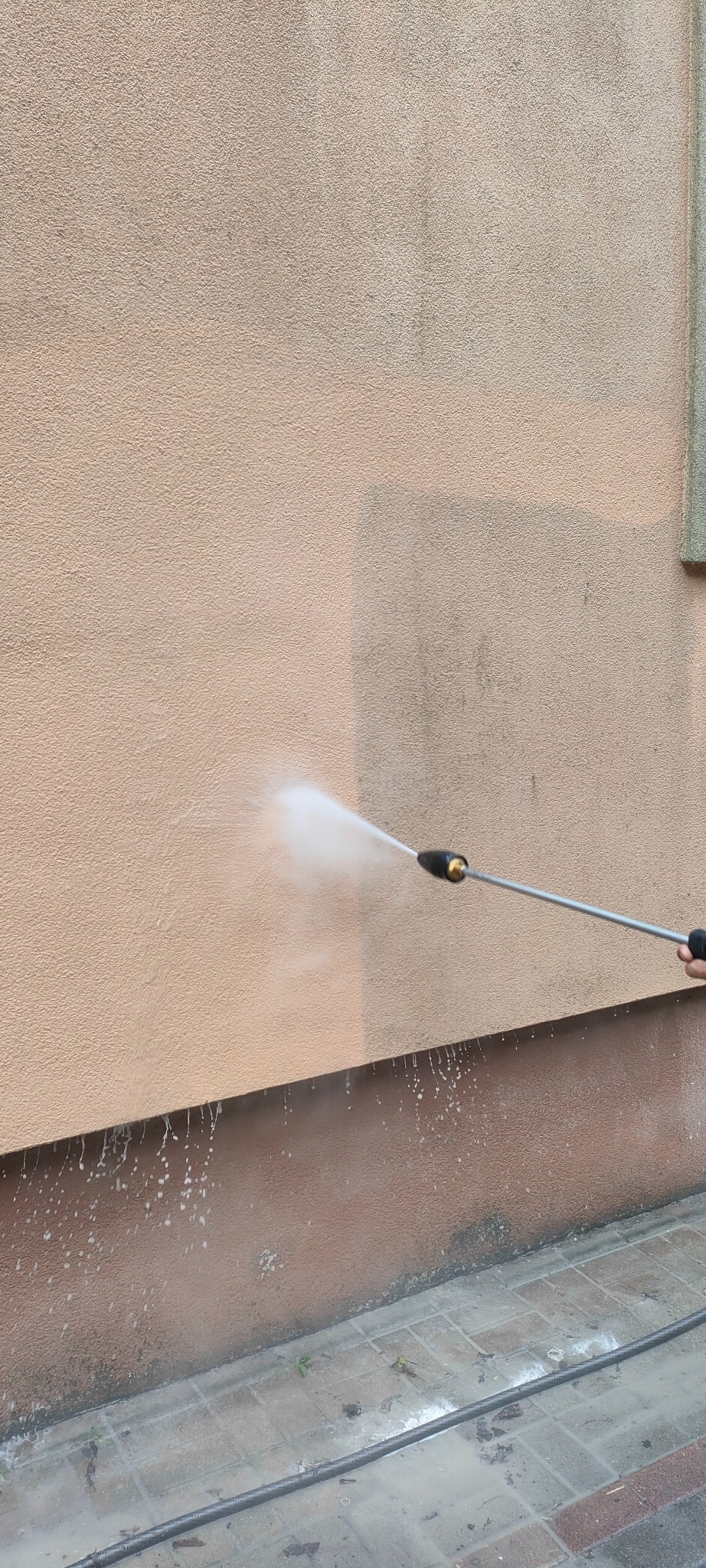 Czyszczenie elewacji budynku myjką ciśnieniową. Widoczna różnica w kolorze między umytą a brudną częścią ściany. Woda spływa po elewacji i kostce brukowej.