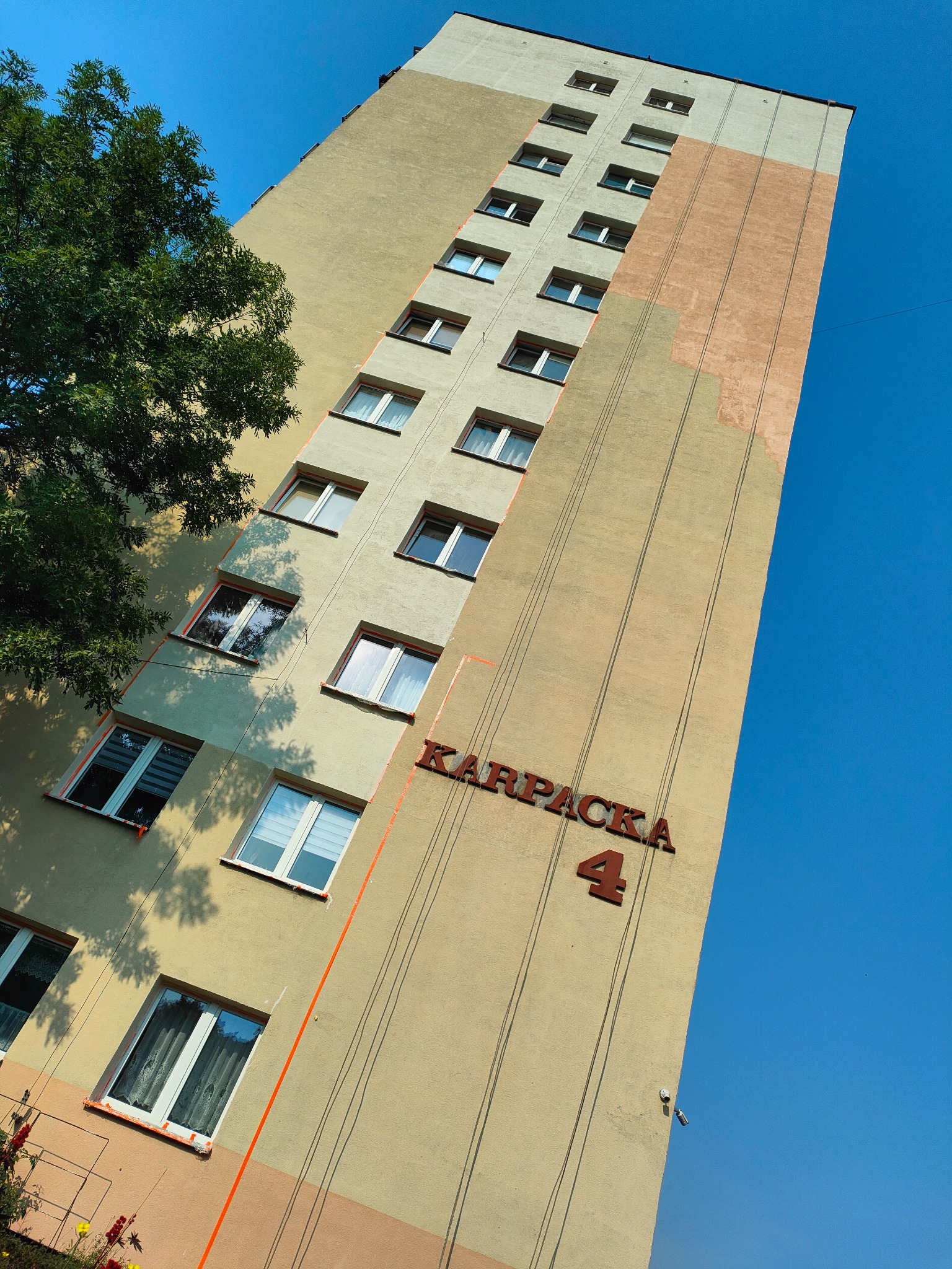 Wysoki blok mieszkalny z częściowo odnowioną elewacją w odcieniach beżu i brązu, widoczny z dołu pod kątem, z napisem 'Karpacka 4' i fragmentem drzewa.