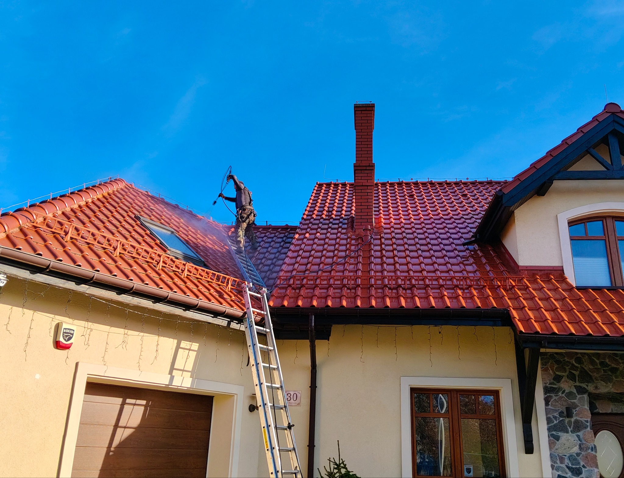 Czyszczenie dachu z czerwonej dachówki myjką ciśnieniową przez pracownika na drabinie. Widoczny budynek z beżową elewacją i błękitne niebo.