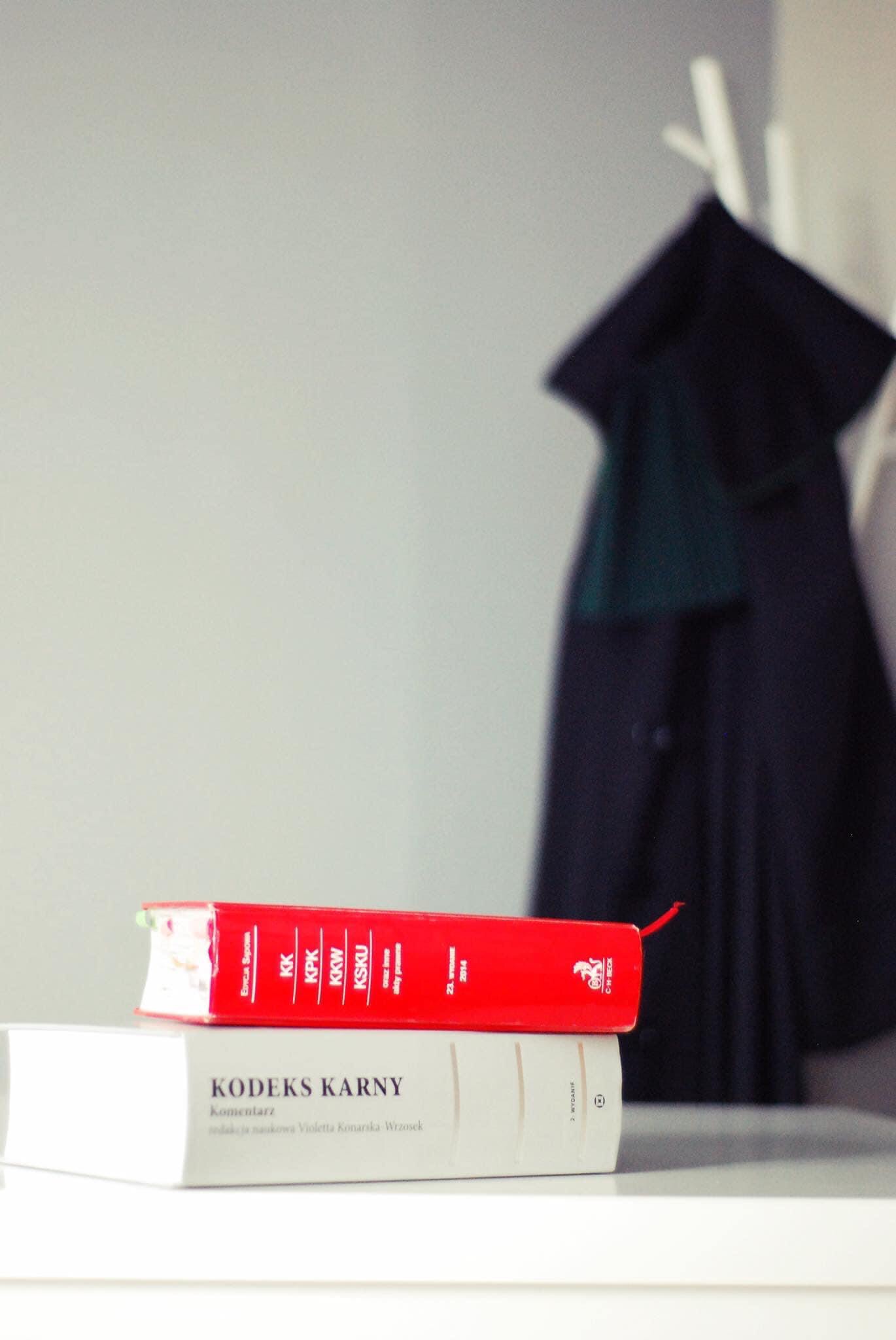 Stos kodeksów prawnych, w tym 'Kodeks Karny', na blacie, z tokiem adwokackim wiszącym w tle na wieszaku.