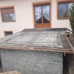 Budowa piwnicy ziemnej z betonowych pustaków, zbrojenie stalowe na wierzchu, widoczny wąż drenażowy w wykopie, dom jednorodzinny w tle.