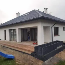 Nowoczesny dom jednorodzinny z jasną elewacją, tarasem z kostki brukowej w trakcie układania, widoczne obrzeża i przygotowane podłoże, w tle niebieska zjeżdżalnia.