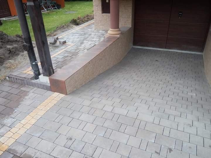 Ułożona szara kostka brukowa przed garażem z brązową bramą i kolumną z dekoracyjną ścianką, z żółtym pasem kostki wzdłuż krawędzi ścieżki.