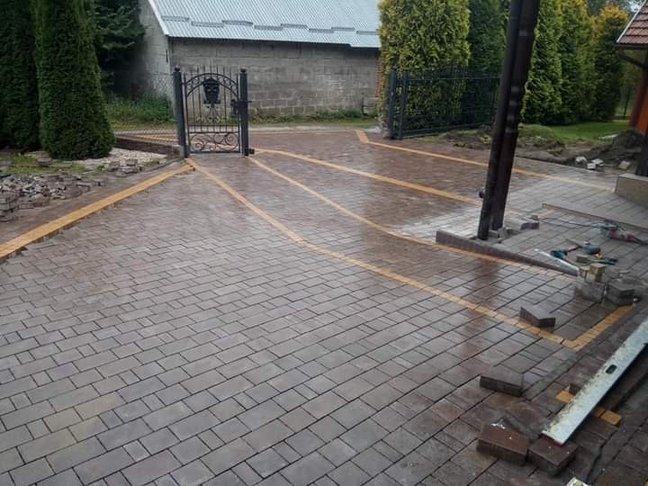 Ułożona, mokra nawierzchnia z szarej kostki brukowej z żółtymi pasami, prowadząca do metalowej bramy wjazdowej i częściowo ułożonych schodów wejściowych do domu.