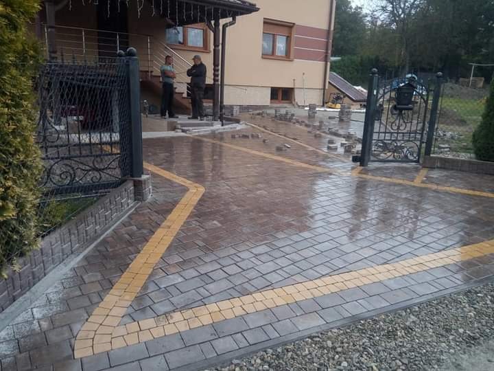 Ułożona kostka brukowa w kolorze szaro-brązowym z żółtymi akcentami tworzącymi obramowanie, częściowo mokra powierzchnia, w tle budynek mieszkalny i dwóch mężczyzn przy wejściu.