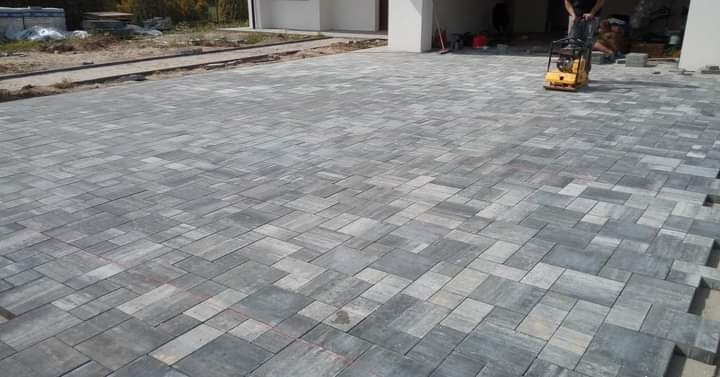 Ułożona szara kostka brukowa na podjeździe, zagęszczarka wibracyjna w użyciu, widoczne krawężniki i niedokończone obrzeże.