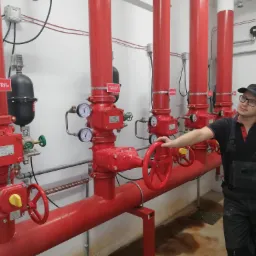 Instalator w kombinezonie roboczym przy czerwonych rurach i zaworach systemu przeciwpożarowego w pomieszczeniu technicznym.