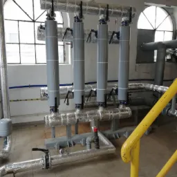 Instalacja grzewcza z czterema pionowymi rurami, izolowanymi i zabezpieczonymi zaworami, w pomieszczeniu z oknami łukowymi i żółtą barierką.