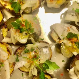 Śledź na pieczonych ziemniakach z cebulą, og&oacute;rkiem kiszonym i zieloną pietruszką, posypany przyprawami, ułożony na metalowej tacy.