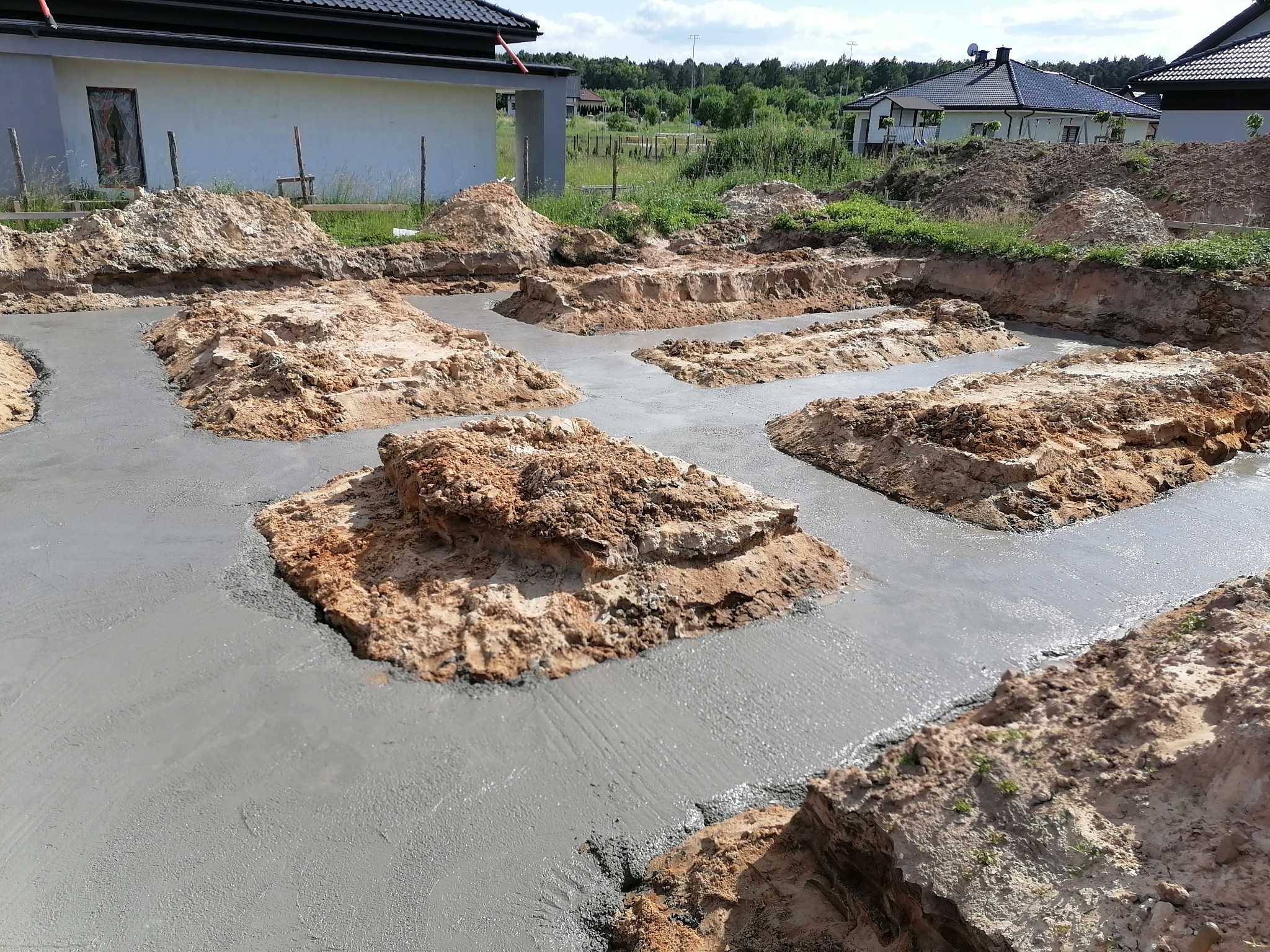 Wylany fundament pod budynek jednorodzinny, widoczne wykopane i zalane betonem ławy fundamentowe, w tle budynek w stanie surowym otwartym.