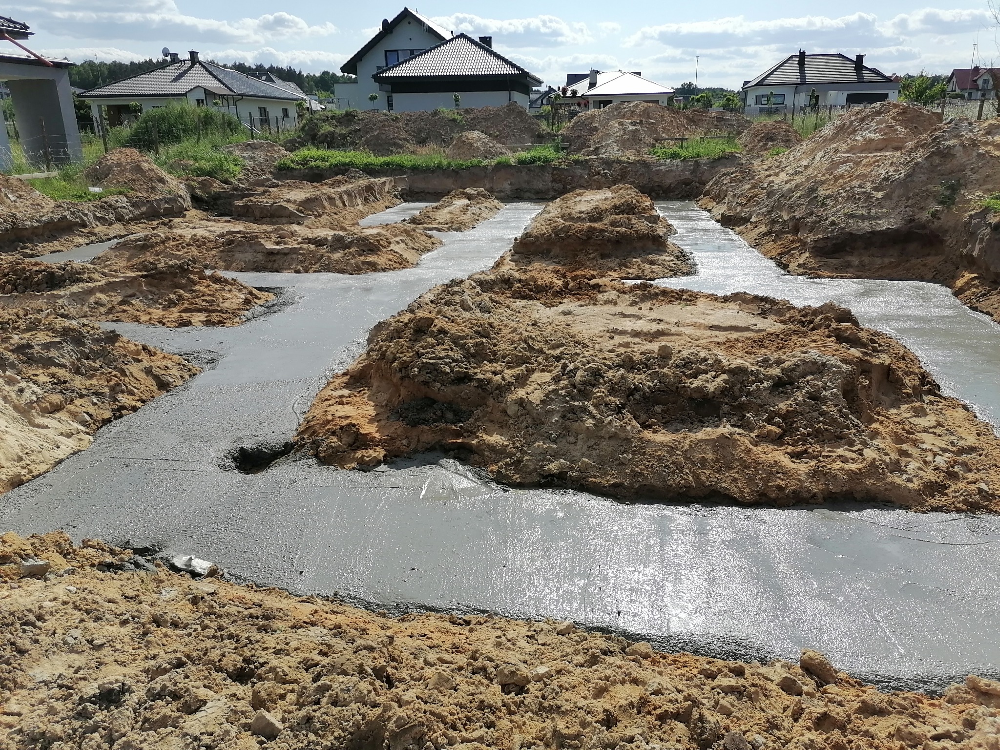 Wylane fundamenty pod dom z widocznymi wykopami i ziemią, w tle domy jednorodzinne pod błękitnym niebem.