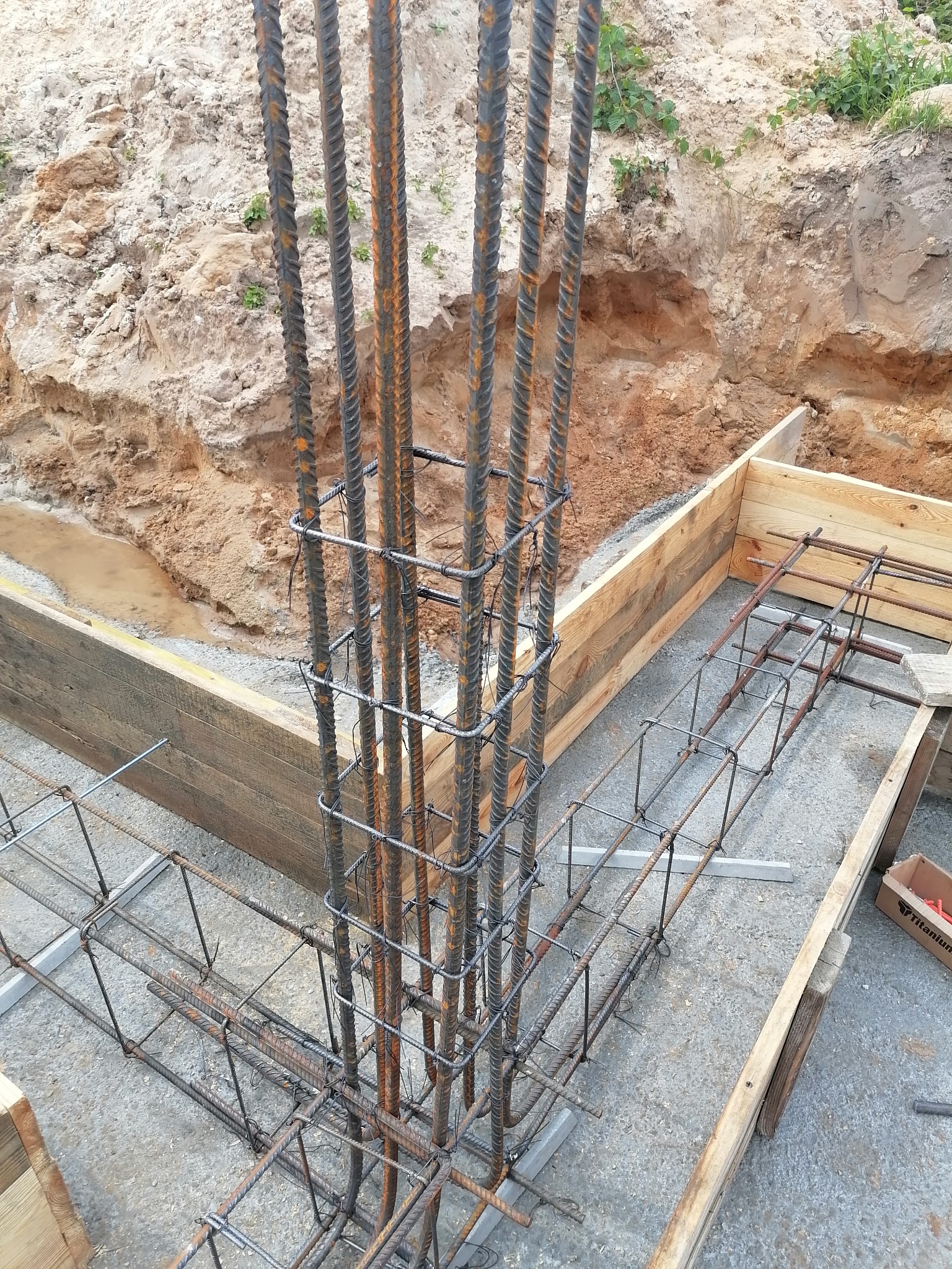 Zbrojenie pionowe i poziome w szalunku fundamentów, widoczny wykop z ziemią i kałużą wody.