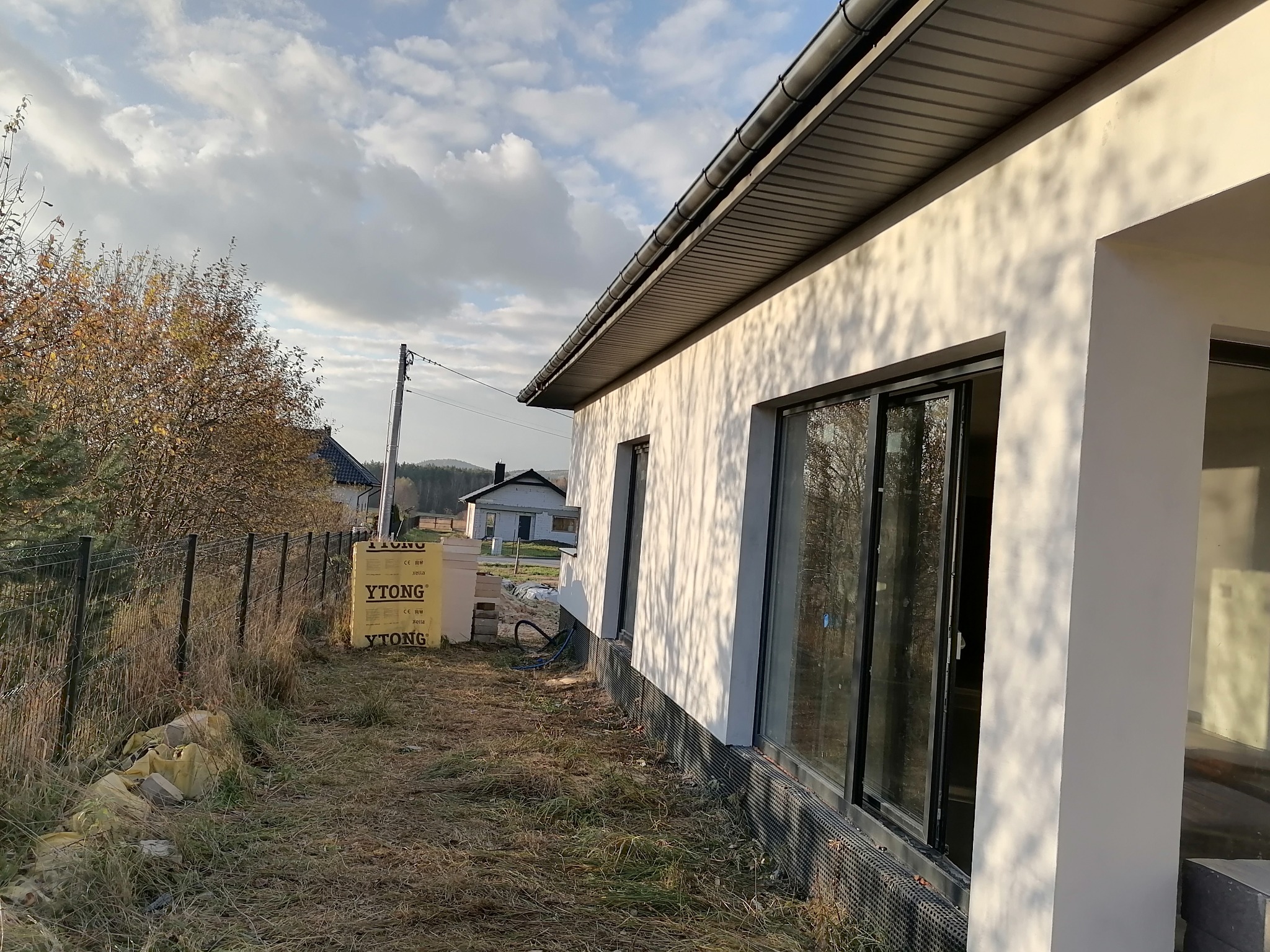 Fragment nowo wybudowanego domu z białym tynkiem, czarnymi oknami i widocznymi bloczkami Ytong na działce.