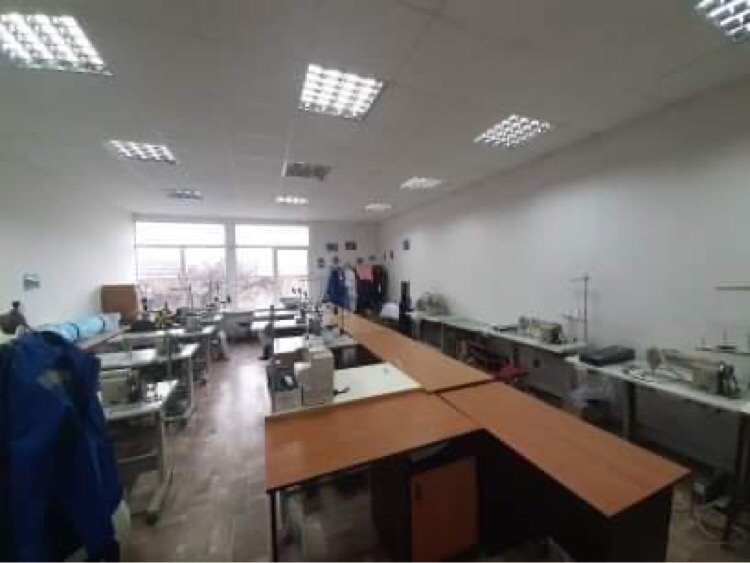 Przestronna pracownia krawiecka z rzędami stołów i maszynami do szycia, oświetlona panelami LED, z ubraniami wiszącymi w tle przy oknie.