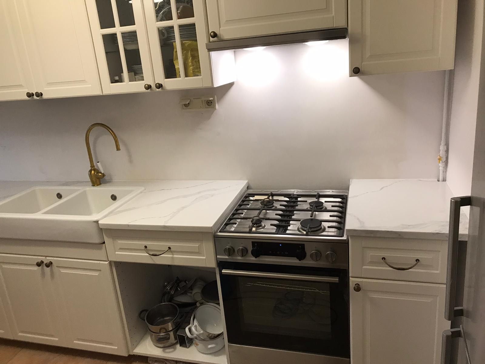 Kuchnia w stylu klasycznym: białe szafki z frezowanymi frontami, złota bateria przy zlewie farmerskim i blat imitujący marmur z szarymi żyłami, obok płyta gazowa.