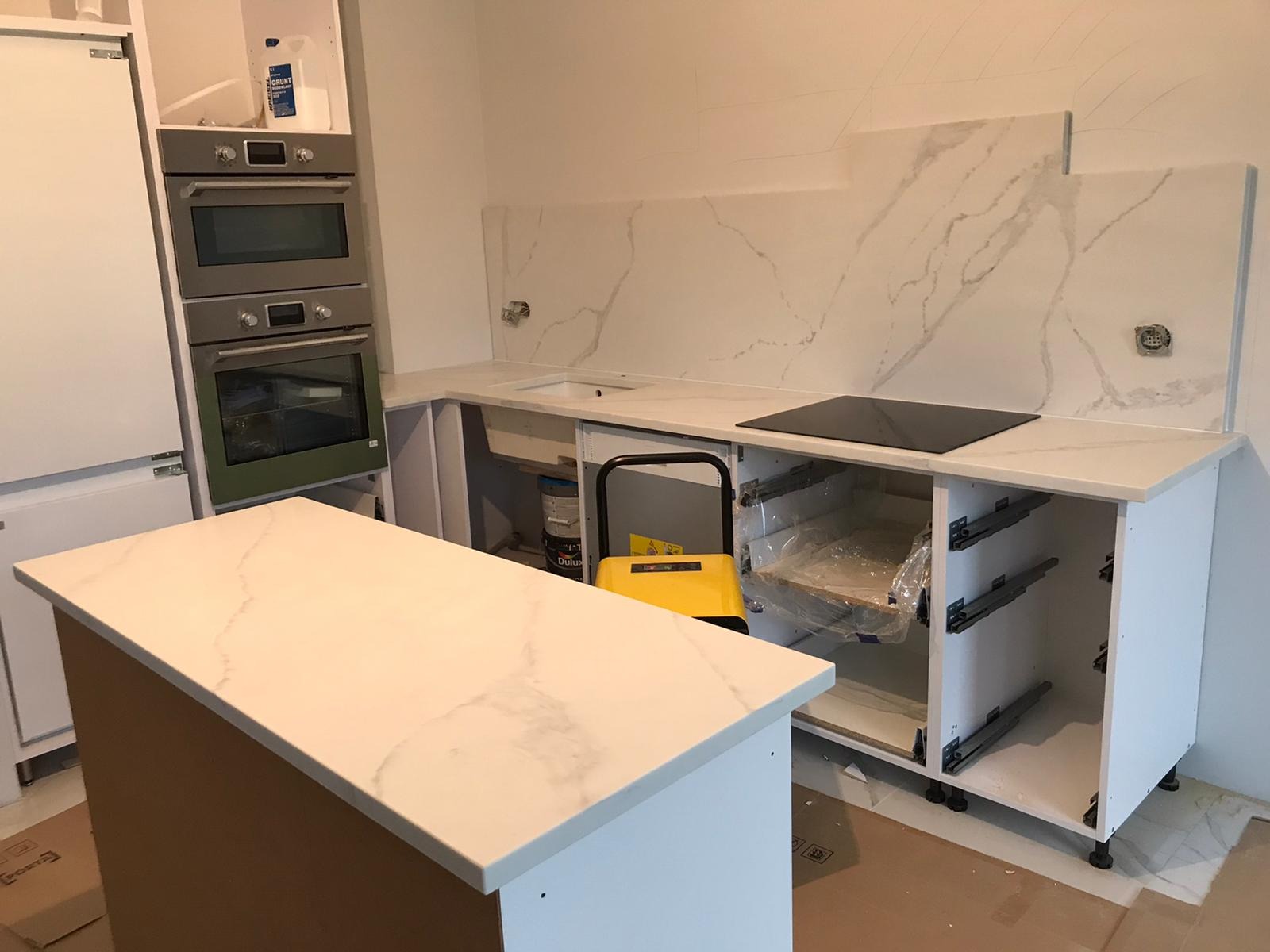 Montaż blatu kamiennego w nowej kuchni, widoczna wyspa kuchenna, płyta indukcyjna i fragment ściany z marmurowym wzorem, etap prac wykończeniowych.