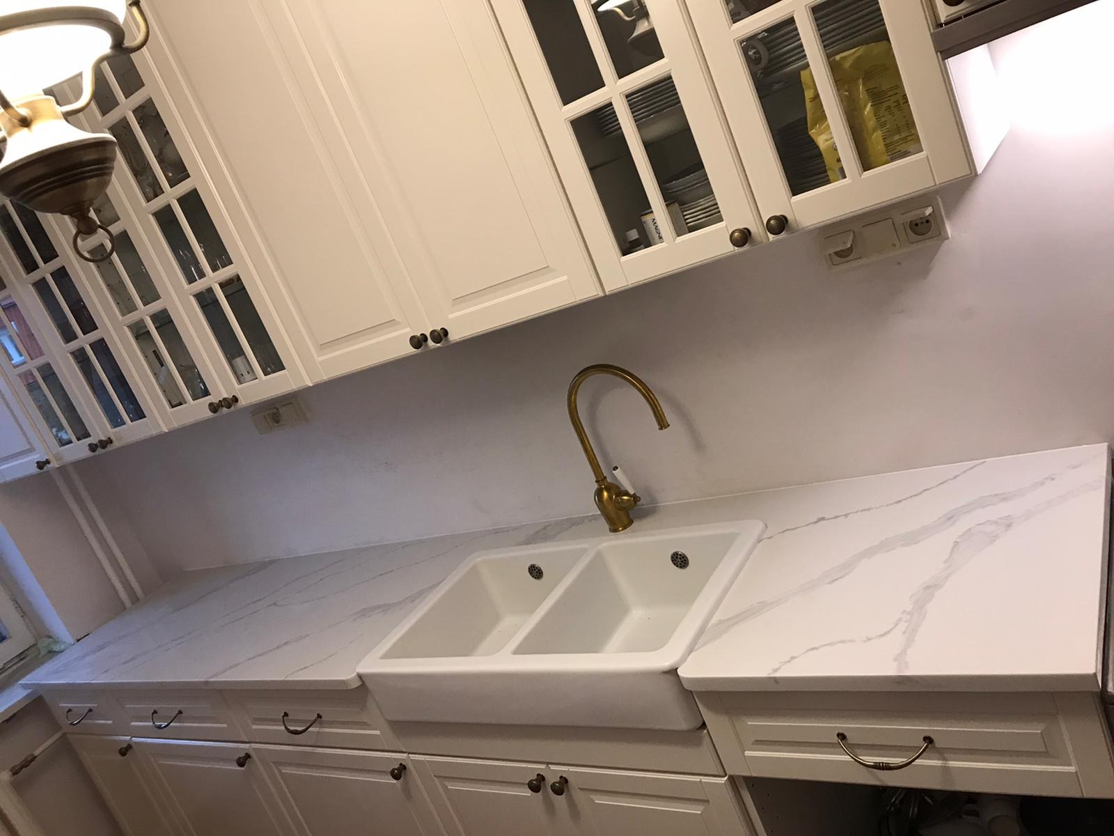 Biała kuchnia w stylu rustykalnym z blatem imitującym marmur, dwukomorowy ceramiczny zlew i mosiężna bateria.