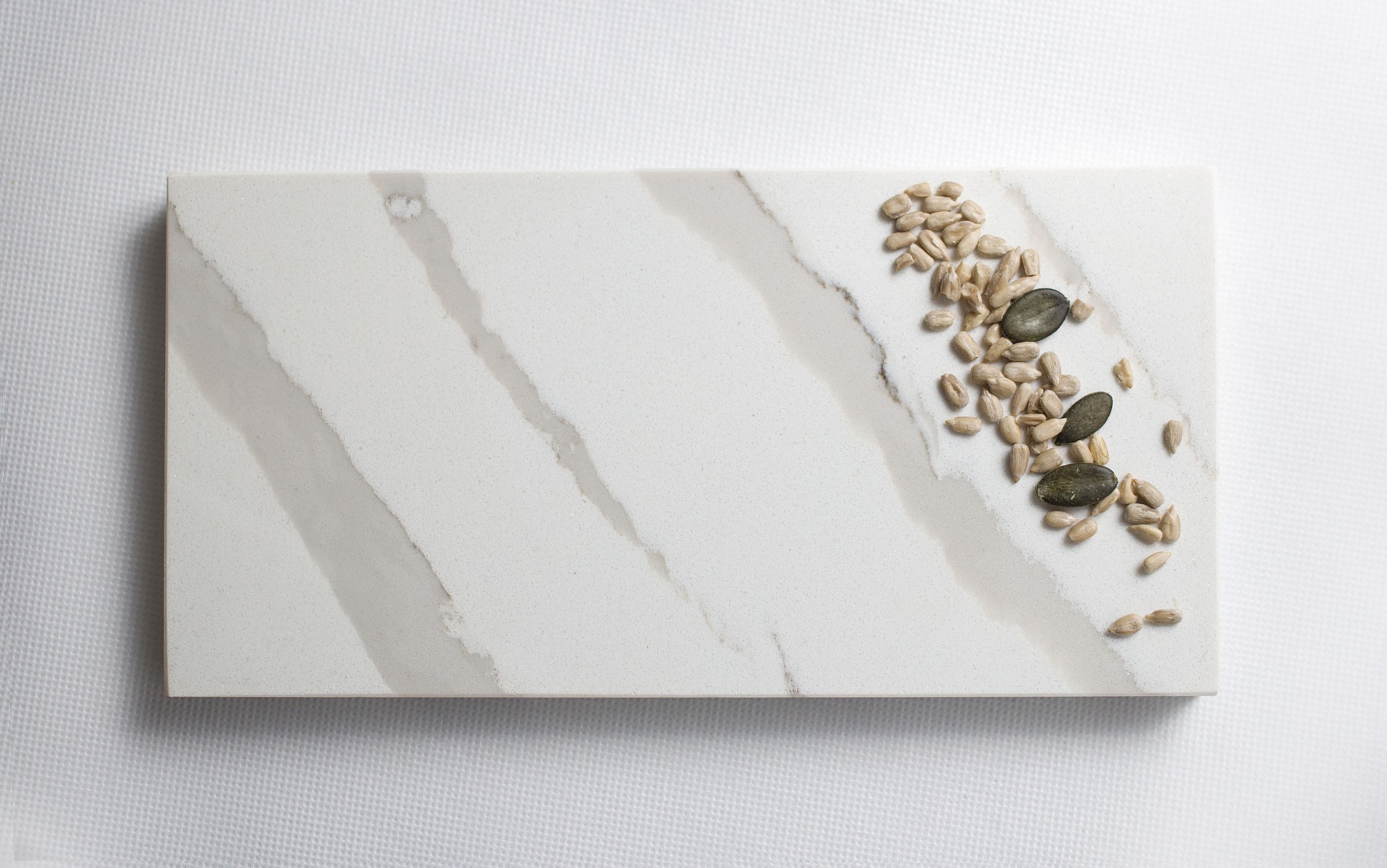 Prostokątny fragment blatu z jasnego konglomeratu z szarymi żyłami, na białym tle z drobnymi nasionami słonecznika i dyni na krawędzi.