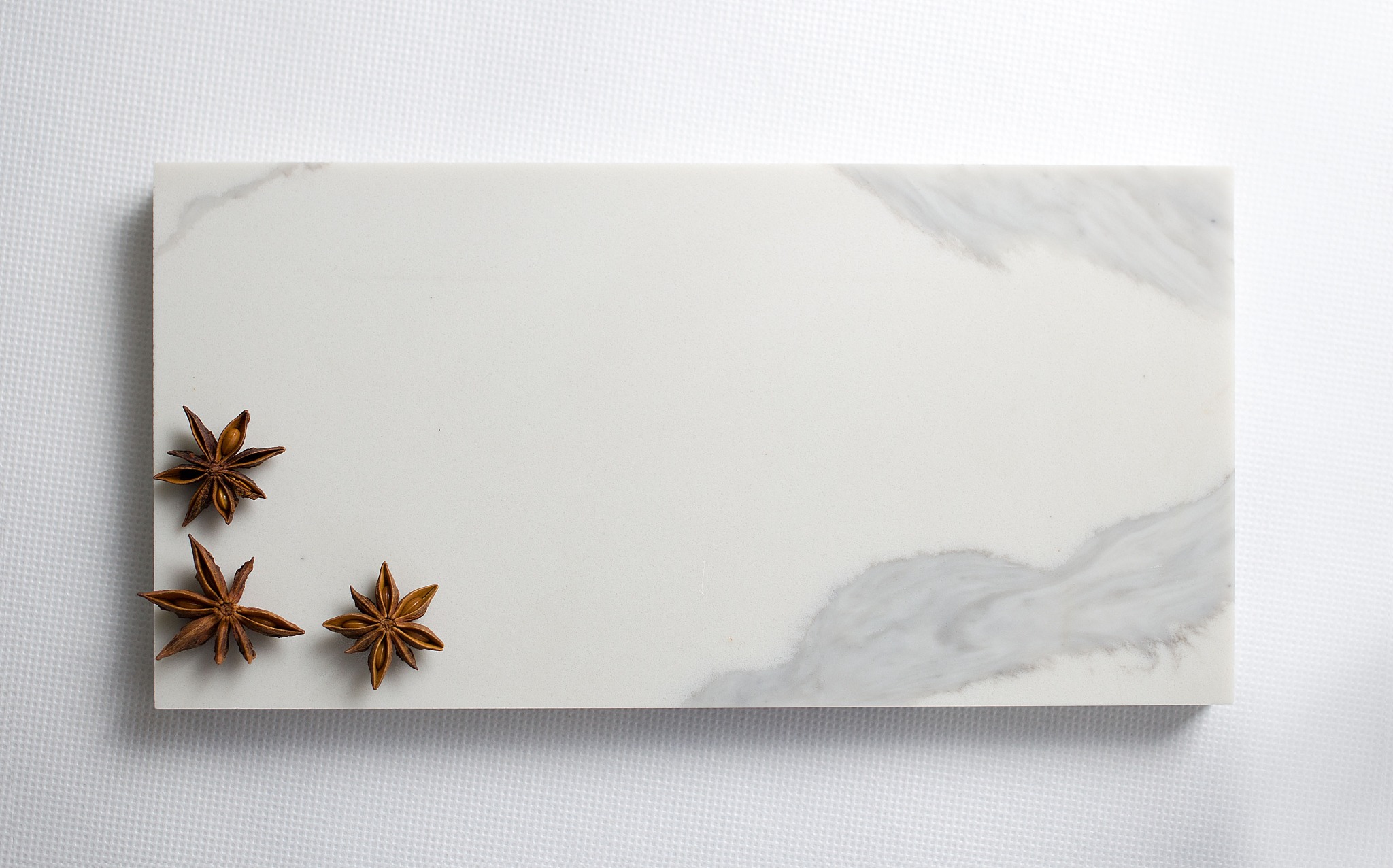 Biały, prostokątny blat z delikatnym szarym wzorem marmuru, ozdobiony trzema gwiazdkami anyżu w lewym dolnym rogu, leżący na białej teksturowanej powierzchni.