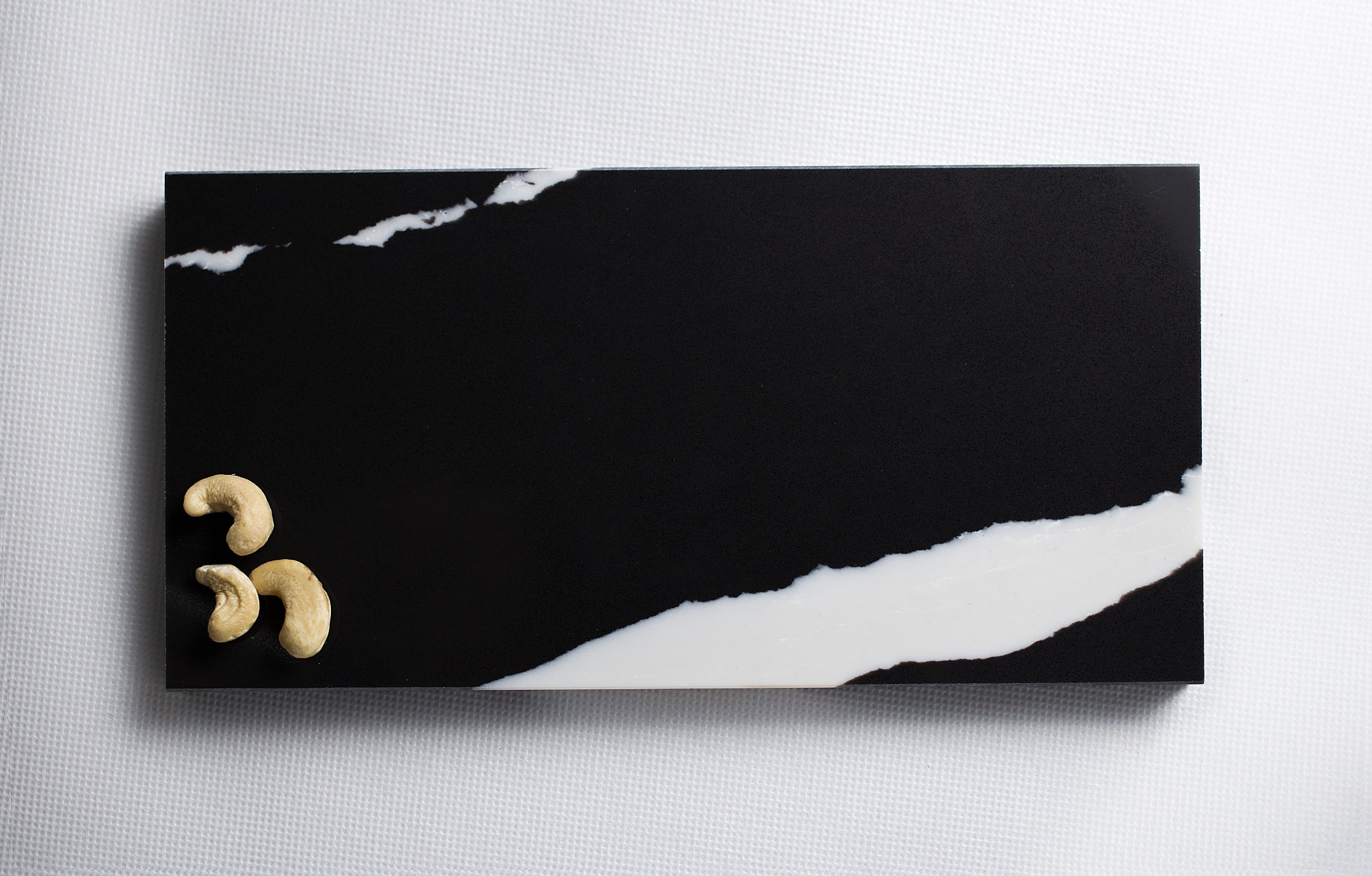 Czarny prostokątny element dekoracyjny z białym żyłkowaniem, ułożony na białej tekstylnej powierzchni, z trzema orzechami nerkowca w lewym dolnym rogu.