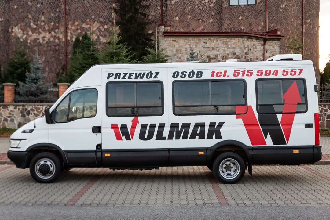 Biały bus z napisem 'Przewóz osób' i numerem telefonu, logo firmy WULMAK z czerwoną strzałką, zaparkowany na chodniku z kostki brukowej, na tle kamiennej ściany budynku.