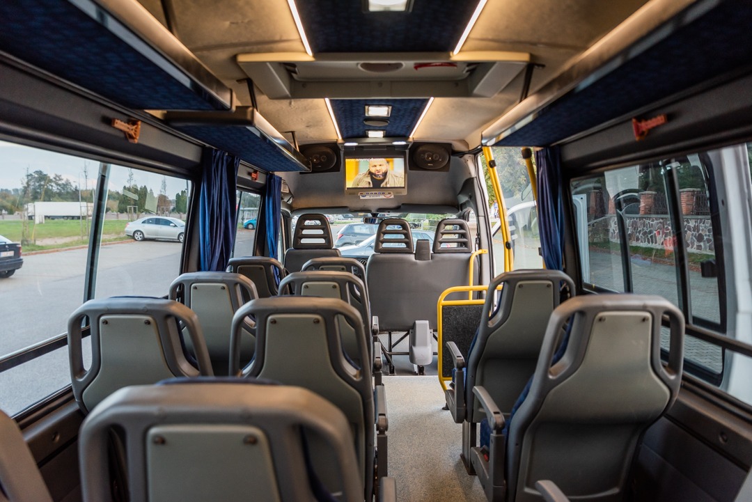 Wnętrze autobusu z rzędami szarych siedzeń, ekranem z włączonym programem telewizyjnym i niebieskimi zasłonami przy oknach.
