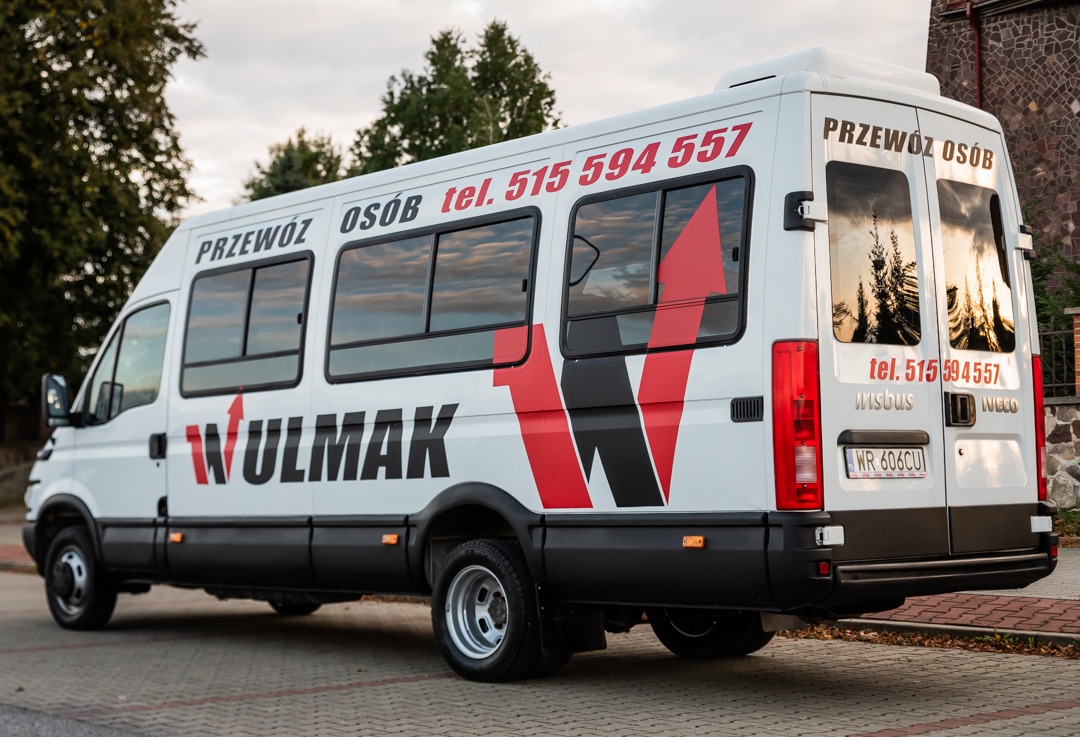 Biały bus z napisem WULMAK i numerem telefonu 515 594 557, zaparkowany na kostce brukowej, widok z tyłu i boku, z drzewami i budynkiem w tle.