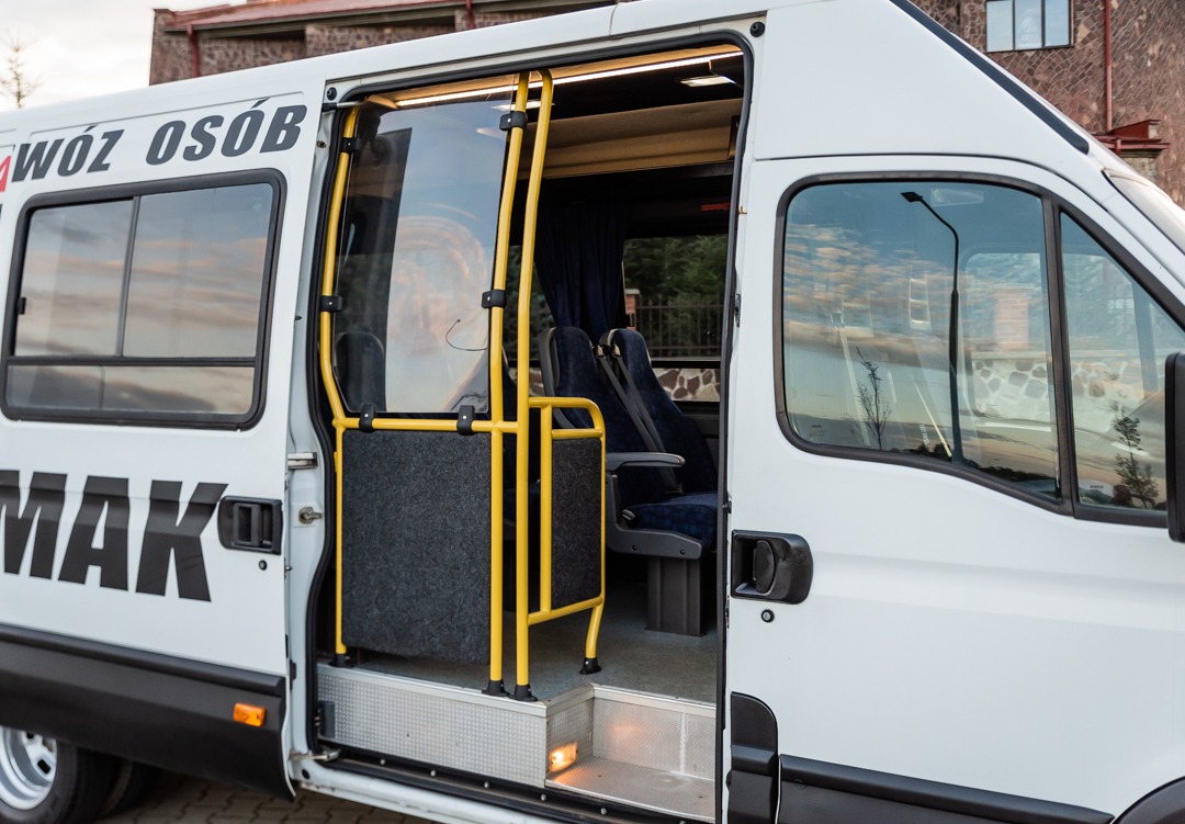 Biały bus z otwartymi przesuwnymi drzwiami, widoczne wnętrze z niebieskimi fotelami i żółtymi poręczami, napis 'WÓZ OSÓB' i nazwa firmy na boku pojazdu.
