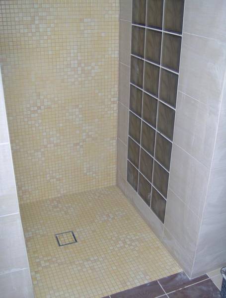 Wykończony prysznic z mozaiką w odcieniach beżu i brązu, szklane luksfery tworzą ściankę działową, widoczny odpływ liniowy w podłodze.