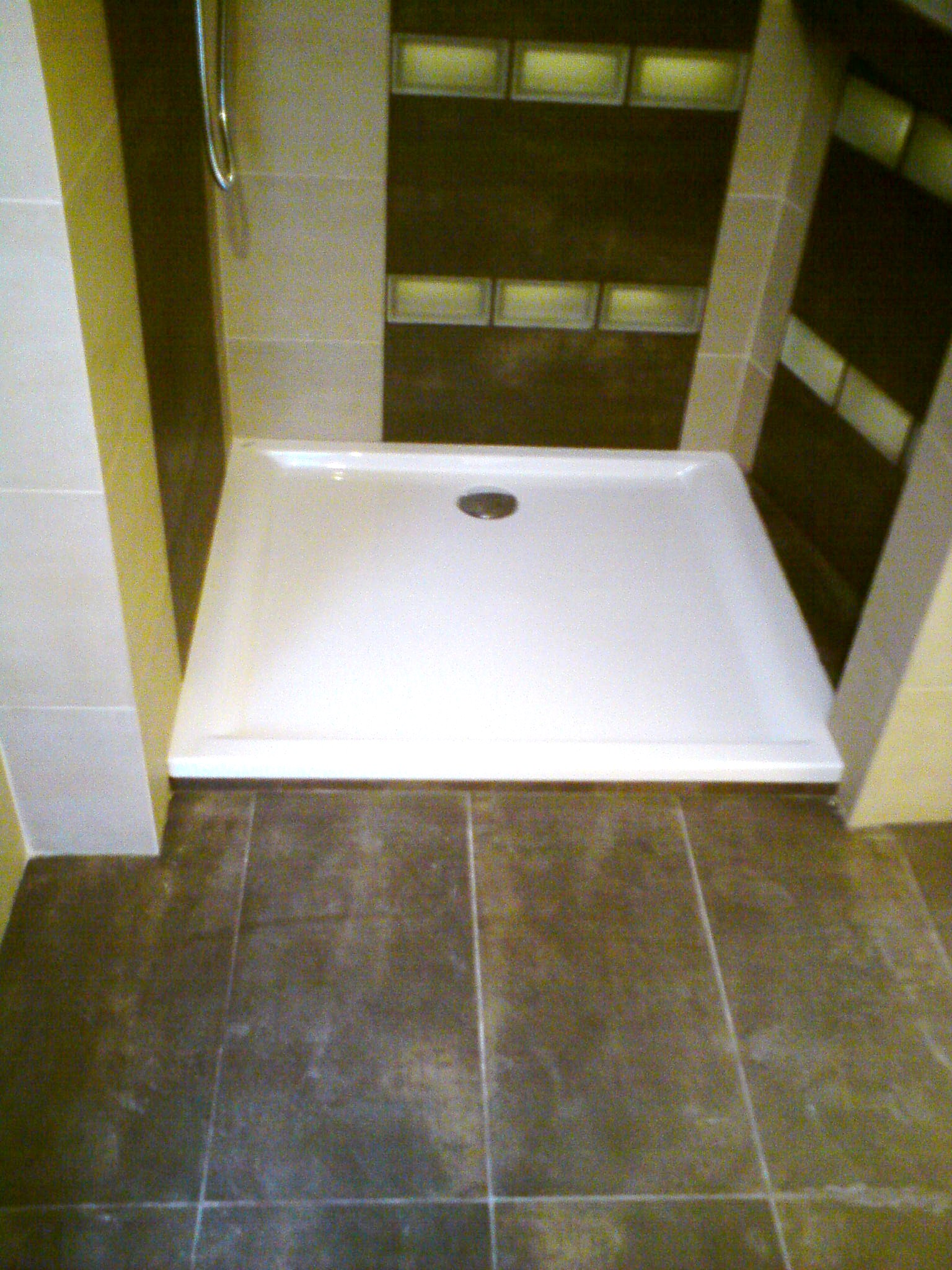 Biały brodzik prostokątny z odpływem centralnym, osadzony w brązowej, kwadratowej płytce podłogowej, otoczony beżowymi płytkami ściennymi i fragmentem panelu z luksferami w tle.