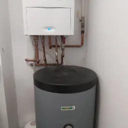 Biały piec centralnego ogrzewania z niebieską diodą i szary zasobnik wody z manometrem, połączone miedzianymi rurami w rogu pomieszczenia.