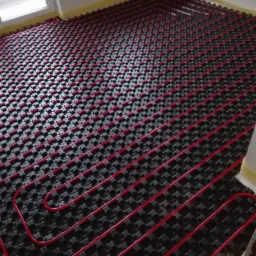 Czerwone rury układane w równych odstępach na czarnej macie pod system ogrzewania podłogowego, widok z góry na etapie instalacji.