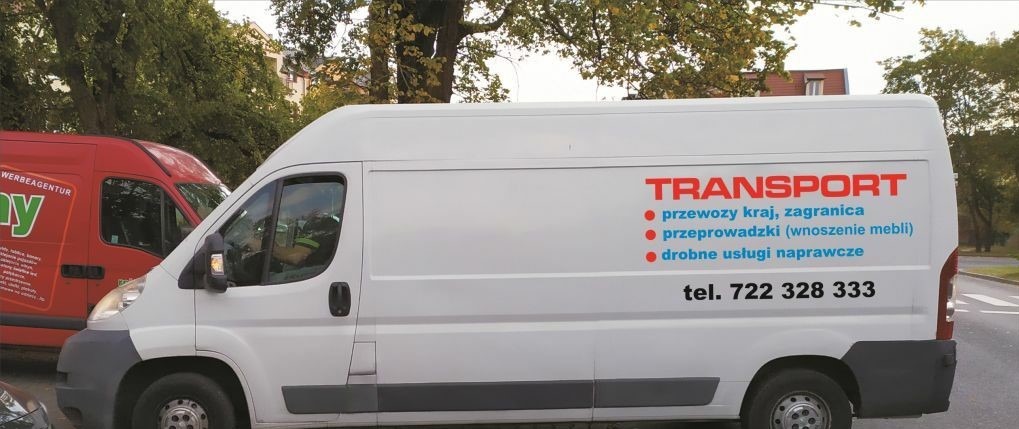 Biały bus z czerwonym napisem TRANSPORT, oferujący przewozy krajowe, zagraniczne, przeprowadzki z wnoszeniem mebli oraz drobne usługi naprawcze, widoczny numer telefonu kontaktowego, na tle zieleni...