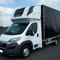 Biały samochód dostawczy Fiat Ducato z czarną plandeką, wyposażony w kabinę sypialną Lamar, zaparkowany na asfaltowym placu w słoneczny dzień.