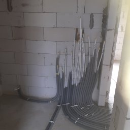 Euromontaż - Szare rury instalacyjne prowadzone wzdłuż ściany z betonu komórkowego, zabezpieczone białymi opaskami zaciskowymi, z wystającymi białymi rurkami oznaczonymi numerami, w surowym stanie deweloperskim.