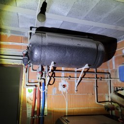 Euromontaż - Szary, poziomy zbiornik akumulacyjny ciepła zawieszony na wspornikach w surowym pomieszczeniu z ceglanymi ścianami i betonowym sufitem, widoczne rury, armatura i wisząca żarówka.