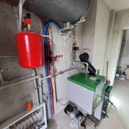 Euromontaż - Zainstalowana kotłownia z zielonym kotłem, czerwonym zbiornikiem wyrównawczym i szarym zasobnikiem, widoczne rury instalacyjne w kolorach niebieskim i czerwonym, surowe ściany w tle.