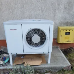 Biała pompa ciepła Stiebel Eltron zamontowana na betonowym fundamencie przy ścianie budynku, obok żółta skrzynka gazowa. W tle widoczne fioletowe kwiaty lawendy i zwinięty przedłużacz.