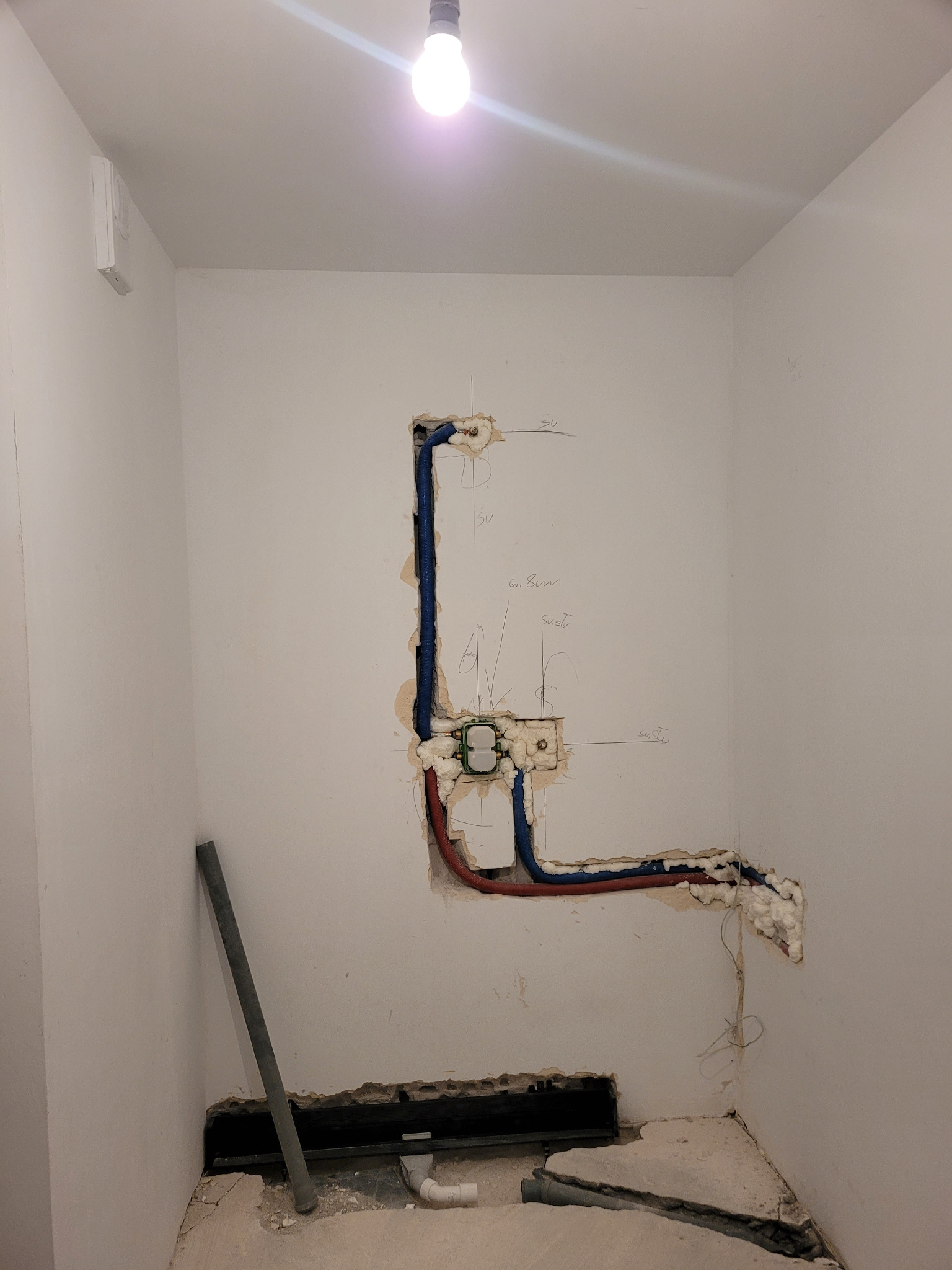 Wnętrze w trakcie prac hydraulicznych: widoczne w ścianie rury w izolacji, otwór w podłodze z fragmentami rur, białe ściany z odręcznymi notatkami i surowym wykończeniem.