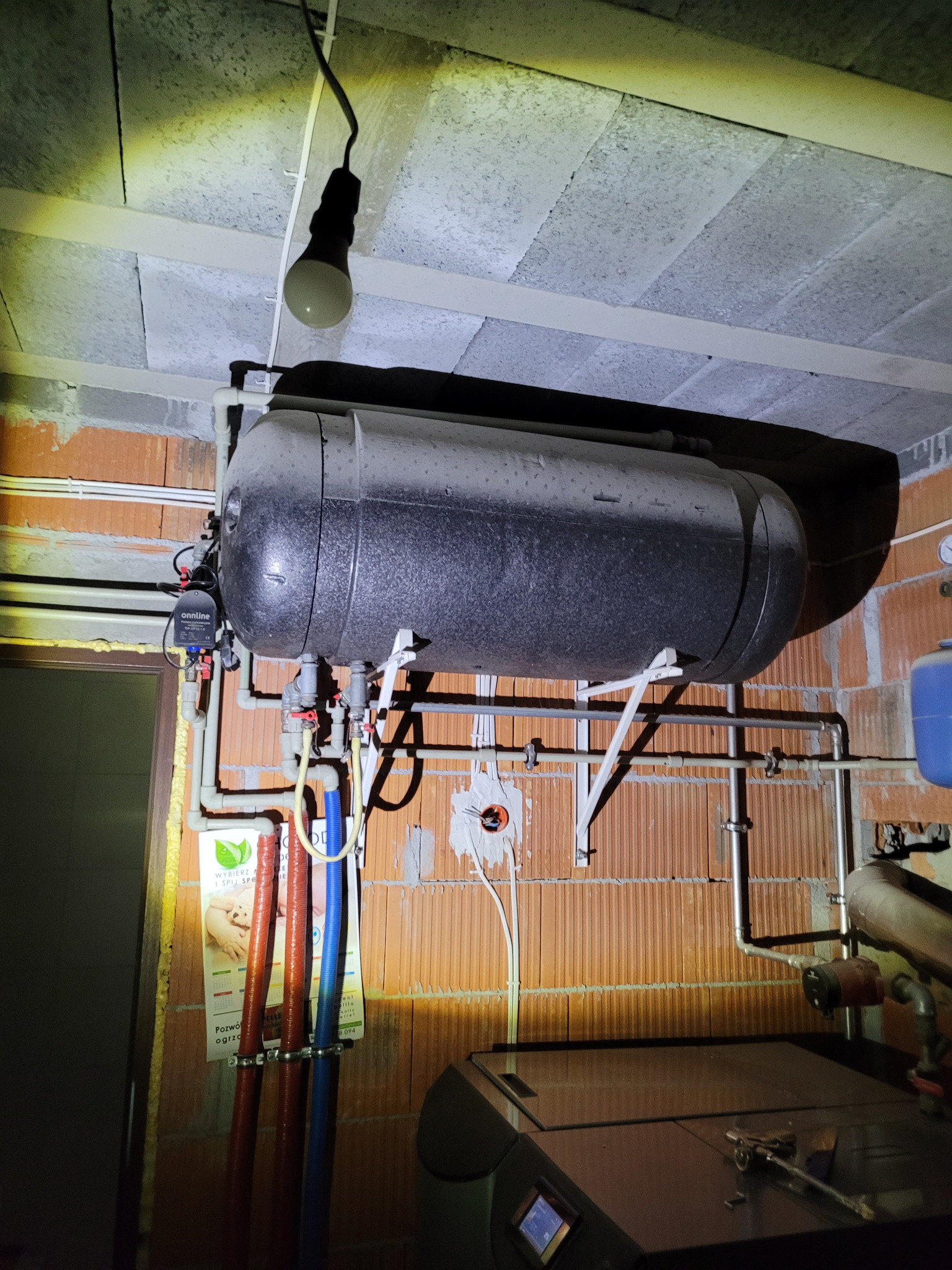 Szary, poziomy zbiornik akumulacyjny ciepła zawieszony na wspornikach w surowym pomieszczeniu z ceglanymi ścianami i betonowym sufitem, widoczne rury, armatura i wisząca żarówka.