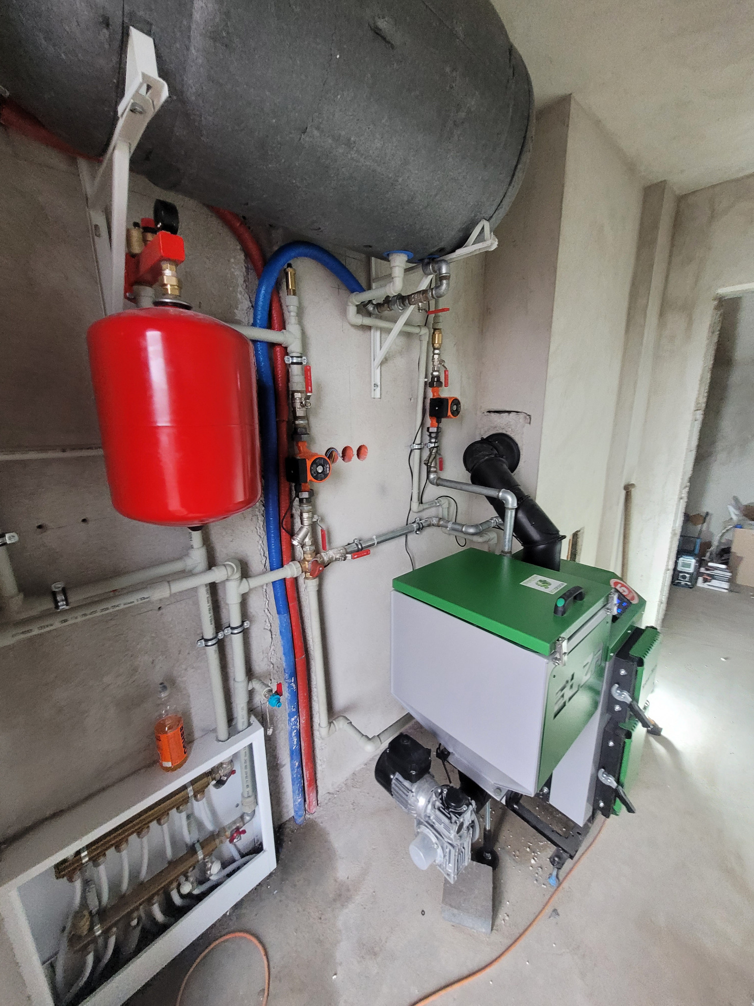 Zainstalowana kotłownia z zielonym kotłem, czerwonym zbiornikiem wyrównawczym i szarym zasobnikiem, widoczne rury instalacyjne w kolorach niebieskim i czerwonym, surowe ściany w tle.