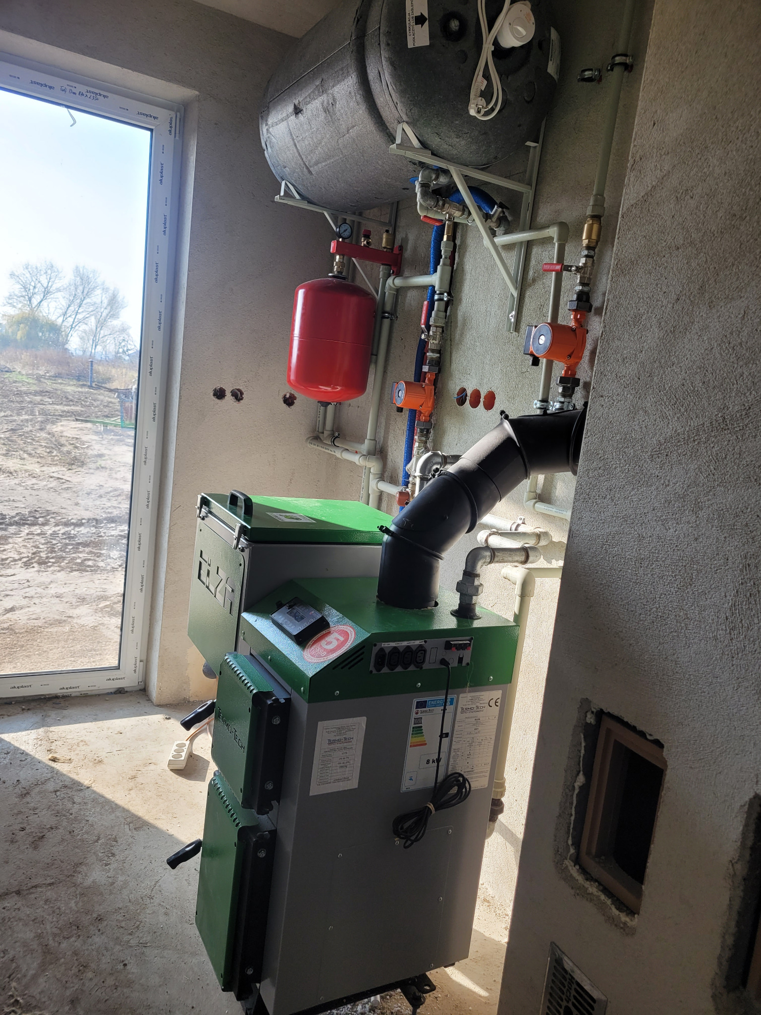 Nowo zainstalowany, zielono-szary piec na paliwo stałe marki Termo-Tech w surowym wnętrzu, z widocznymi rurami, zbiornikiem wyrównawczym i oknem z widokiem na zewnątrz.