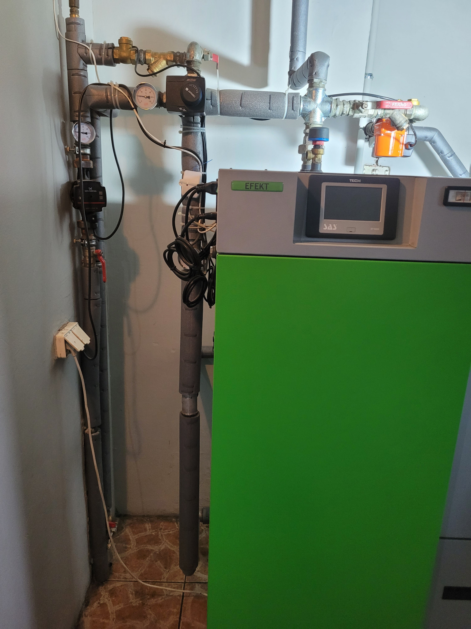 Zielony piec z szarą obudową i wyświetlaczem, podłączony do rur z zaworami i manometrami, widoczne okablowanie, instalacja w pomieszczeniu z brązową podłogą.