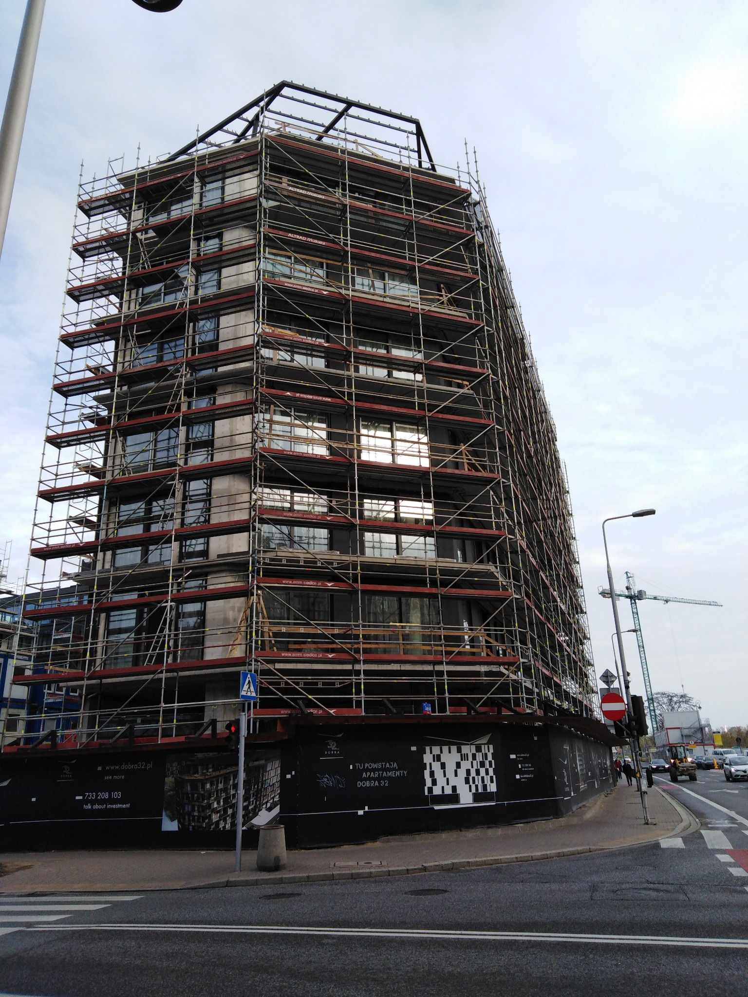 Wielopiętrowy budynek w trakcie budowy, całkowicie otoczony rusztowaniami, widoczny żuraw budowlany w tle, ulica z sygnalizacją świetlną w pierwszym planie.