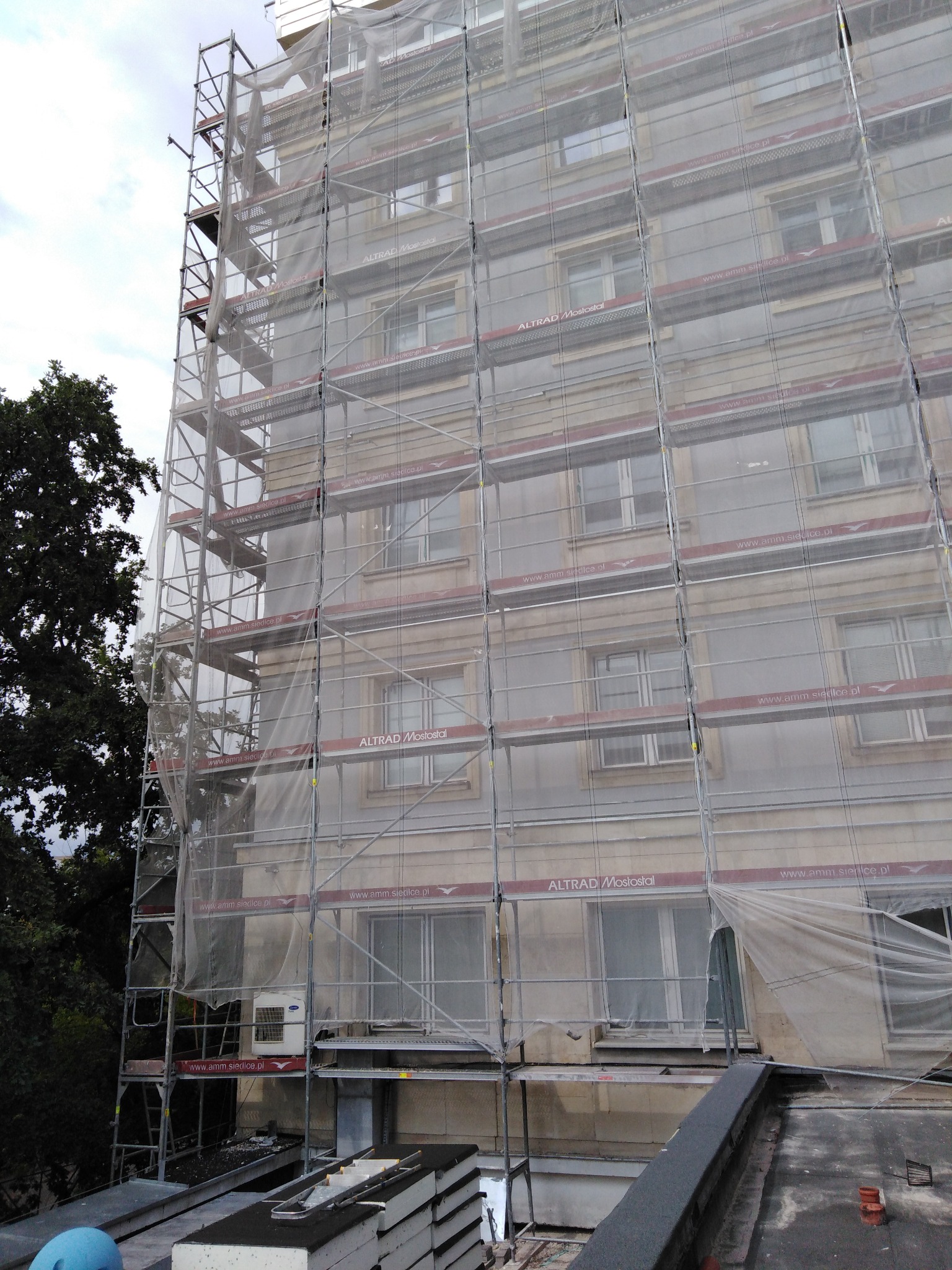 Wielopiętrowy budynek w trakcie remontu, osłonięty siatką ochronną rozpiętą na rusztowaniach. Widoczne okna z ramami, fragment dachu i drzewa w tle.