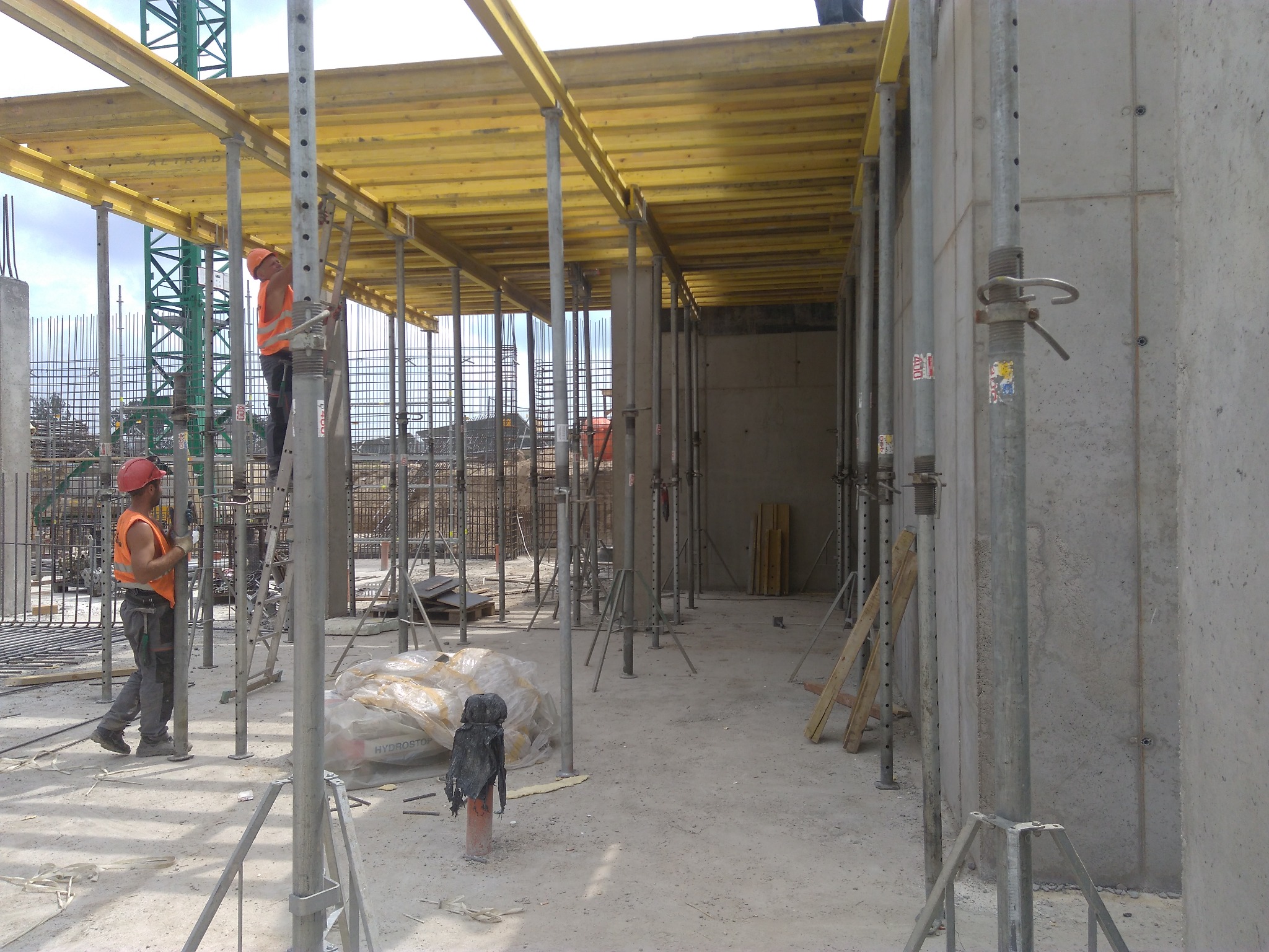 Dwóch pracowników w pomarańczowych kamizelkach na budowie, jeden na drabinie montuje żółte szalunki stropowe, widoczne podpory, betonowe ściany i dźwig w tle.