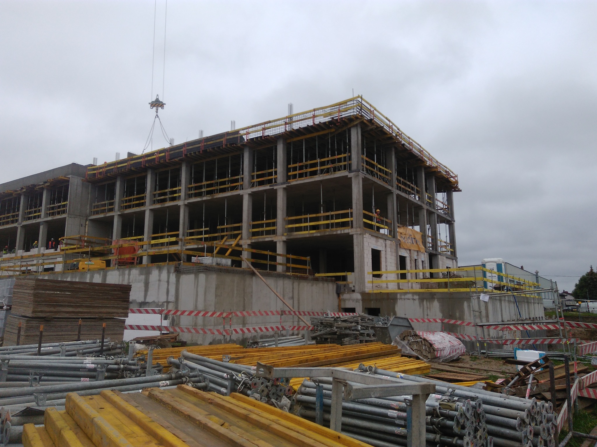 Budowa wielopiętrowego budynku z betonu i stali, widoczne szalunki i rusztowania, dźwig podnosi element konstrukcyjny na tle pochmurnego nieba.