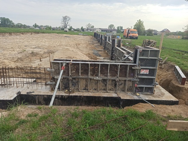 Wykop pod fundamenty z szalunkiem i zbrojeniem, widoczny częściowo zalany beton, w tle teren budowy i samochód dostawczy.