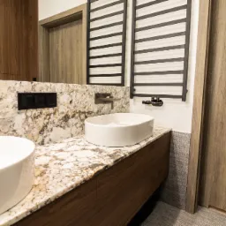 Łazienka z dwoma białymi umywalkami na blacie z kamienia o wzorze przypominającym marmur, nowoczesna armatura w kolorze grafitowym, grzejnik drabinkowy w tle.