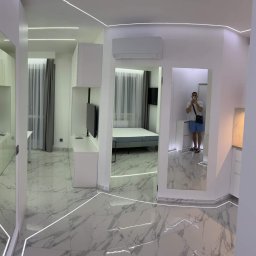 Oleh Tanchak Top Remont - Nowoczesne wnętrze mieszkania w odcieniach bieli z marmurową podłogą, oświetleniem LED wzdłuż ścian i podłogi, oraz odbiciem osoby robiącej zdjęcie w lustrze.